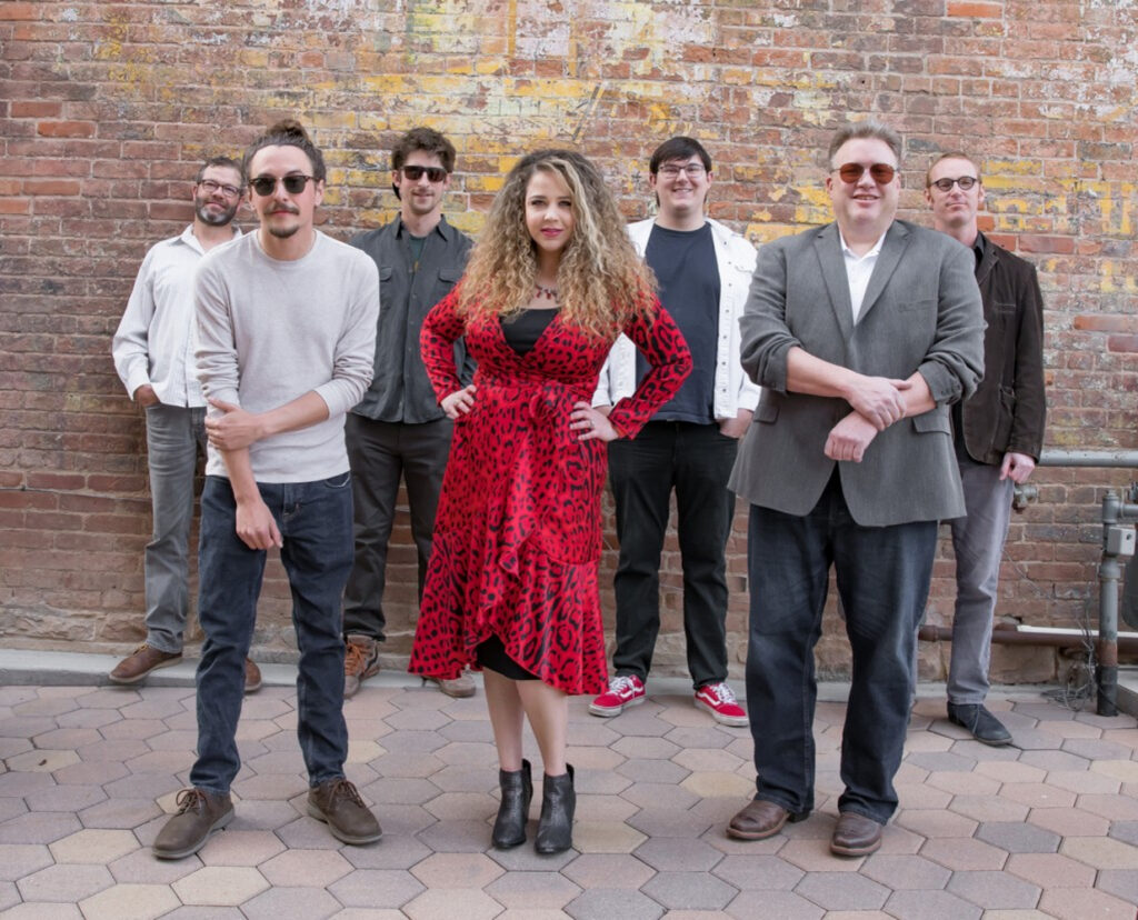 Six bands members plus lead singer Monique pose against brick wall.