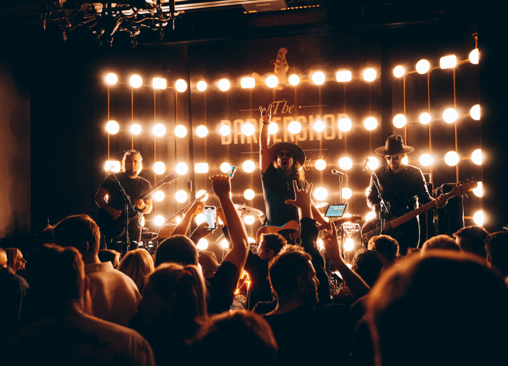 Photo of 4 band members on stage backlit with lightbulbs. Lead singer in the middle playing guitar with hand up.